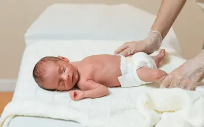 Noworodek śpi na białym kocyku, pielęgniarka w rękawiczkach delikatnie dotyka jego nóżki. Dziecko wygląda na zdrowe, bez oznak hipotrofii.
