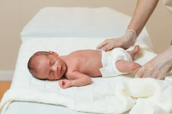 Noworodek śpi na białym kocyku, pielęgniarka w rękawiczkach delikatnie dotyka jego nóżki. Dziecko wygląda na zdrowe, bez oznak hipotrofii.